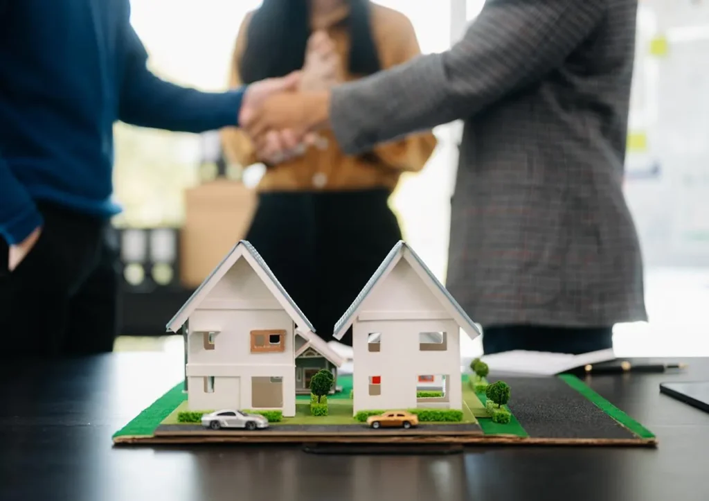 Miniature house models on a table with people shaking hands in the background.