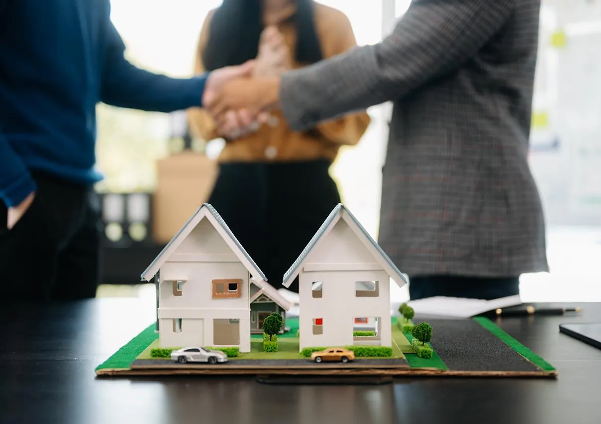 Miniature house models on a table with people shaking hands in the background.